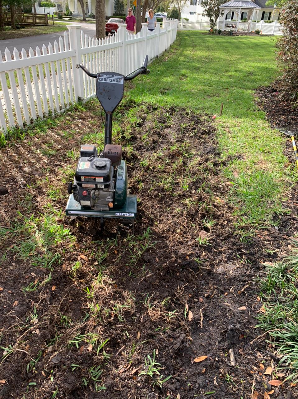 Photo of lawn renewal ground preparation with equipment working the soil near a fence.